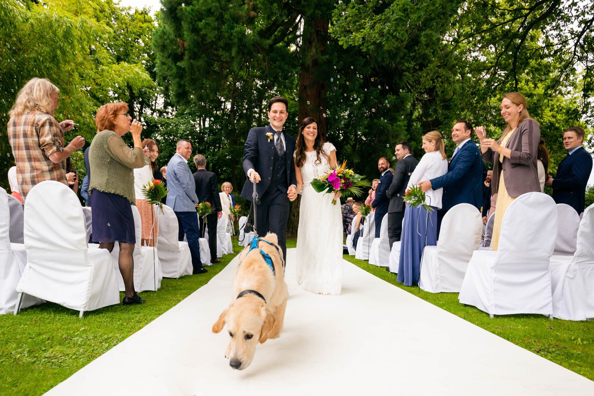 Hochzeitsfoto eines frisch verheirateten Ehepaares, die mit ihrem Hund die Freie Trauung verlassen und an ihren Gästen vorbei zum Sektempfang gehen.