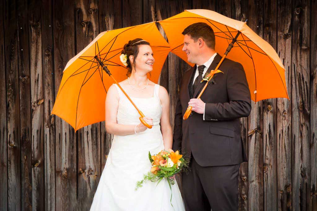 Hochzeitsfotos mit bunten Regenschirmen, Brautpaar mit orangen Schirmen vor Scheunenwand bei Niederschlag